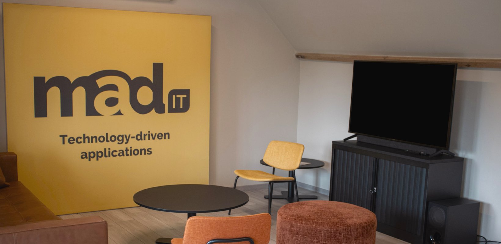 Moderne kantoorruimte van Mad IT met gele wand met logo en slogan "Technology-driven applications", zithoek met designstoelen, ronde tafels en flatscreen-tv op zwart tv-meubel — creatieve werkplek voor technologie en softwareontwikkeling.